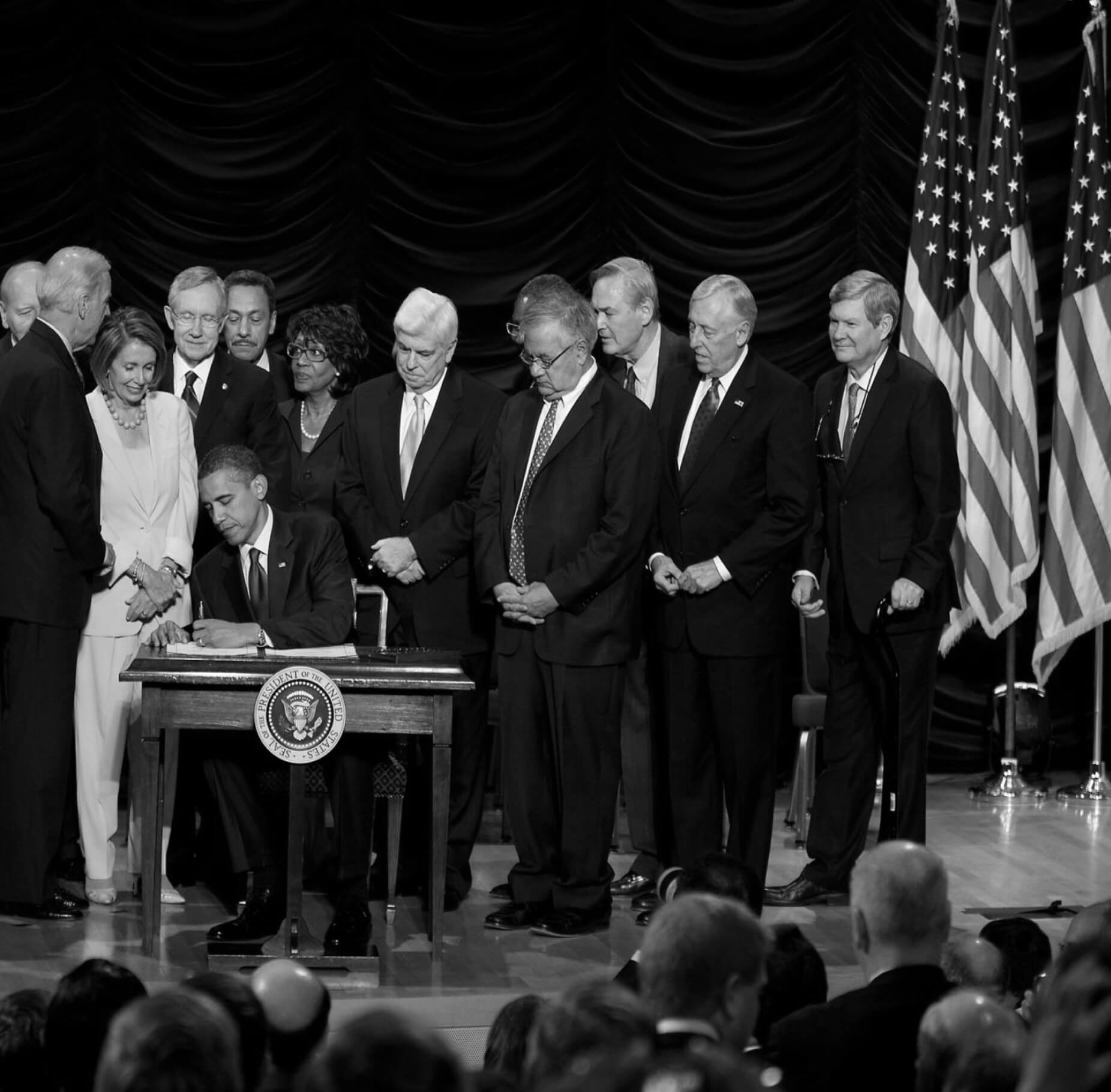 Barack Obama signing a bill with congress behind him.