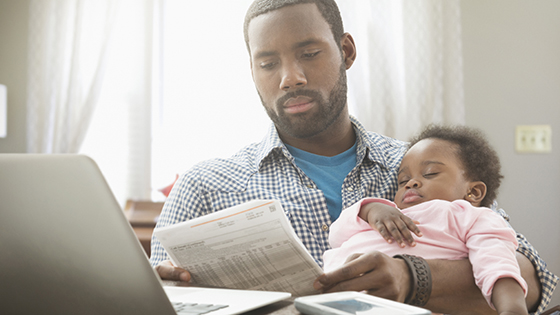 July Article Monthly Image - a young father holding his baby girl in his arms, while reviewing his bills