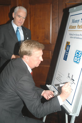 Donald E. Powell, Presidente de la mesa directiva de la FDIC, observa a Gavin Gee, Presidente y CEO de CSBS de 2003, firmar el acuerdo según el cual la CSBS distribuirá y promocionará el programa de estudios para educación financiera Money Smart de la FDIC.