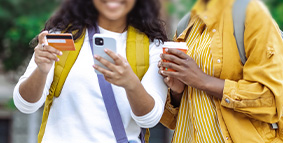 Two young girls, one holding a credit card and cell phone, the other one holding a cup of coffee