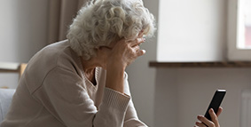 Una anciana sentada en un sofá con una mano en su cabeza mientras sostiene un teléfono inteligente con la otra mano