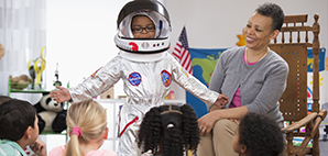 Child wearing astronaut costume in front of clasroom accompanied by teacher
